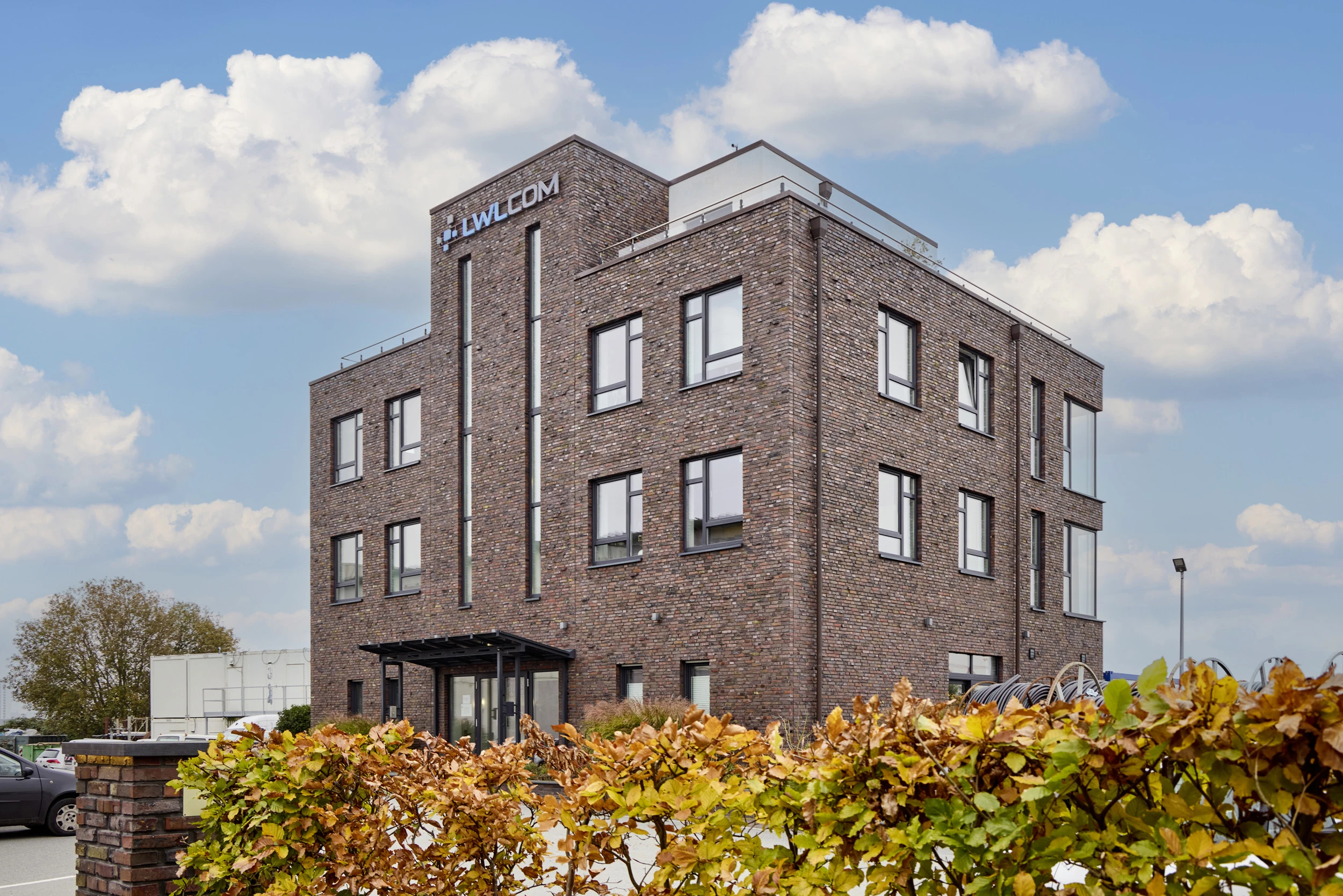 Bürogebäude der LWLcom GmbH aus braun-roten Ziegeln mit mehreren Fenstern, davor eine grün-braun herbstliche Hecke, blauer Himmel mit Wolken.