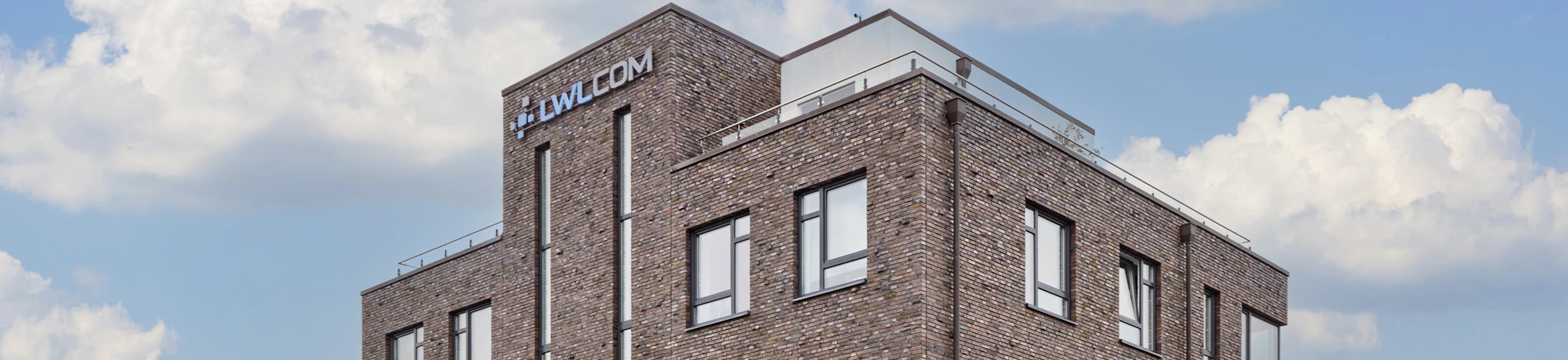 Bürogebäude der LWLcom GmbH aus braun-roten Ziegeln mit mehreren Fenstern, davor eine grün-braun herbstliche Hecke, blauer Himmel mit Wolken.