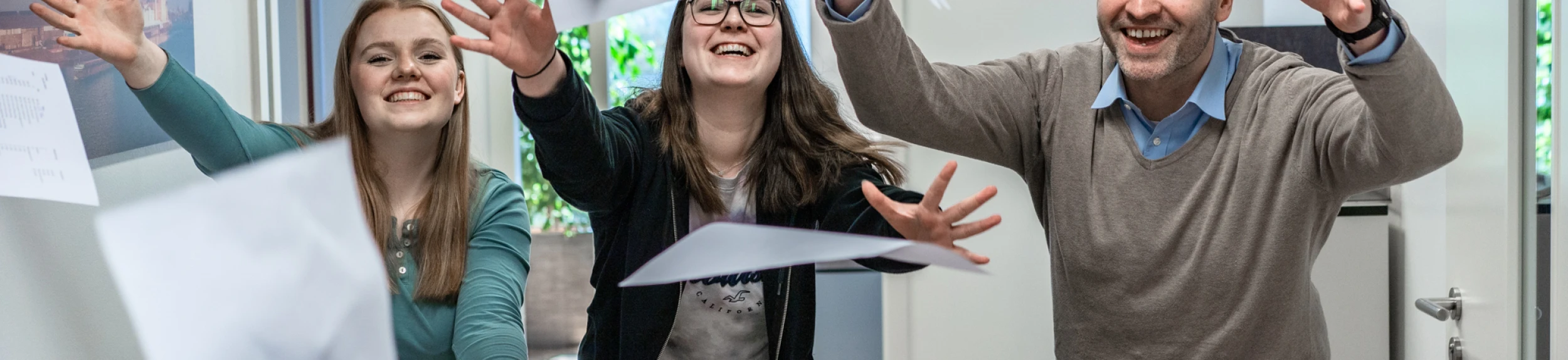 Drei gut gelaunte Mitarbeiter:innen der LWLcom GmbH werfen Papiere in einem Büroflur mit Türen und Fenstern.