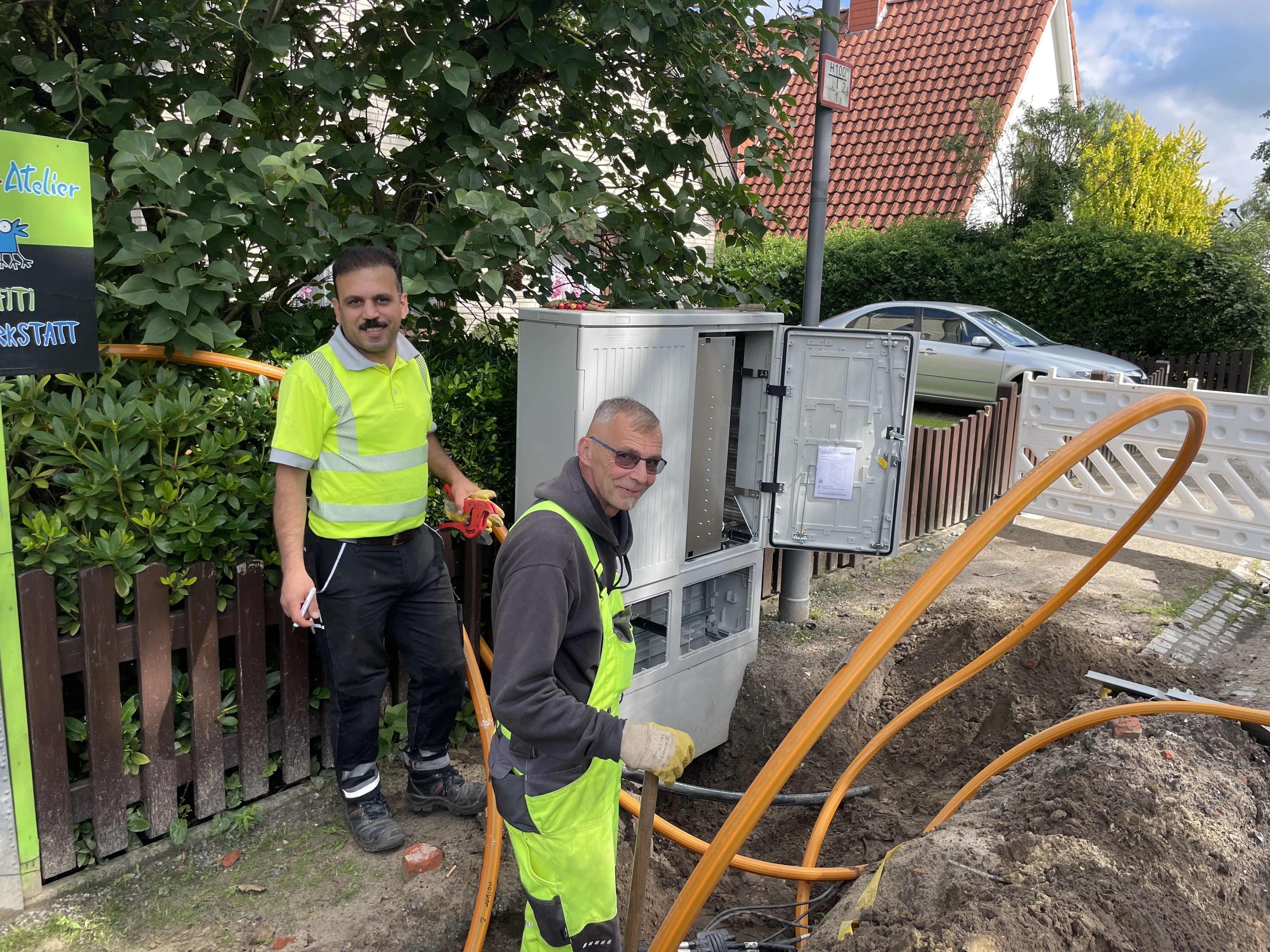 Zwei Bauarbeiter in Warnwesten stehen an einem offenen Technikverteilerkasten am Straßenrand. Mehreren orangefarbene Kabelrohre, die aus dem Boden führen. Im Hintergrund sind ein Gartenzaun, Sträucher und ein Wohnhaus mit rotem Dach zu sehen.