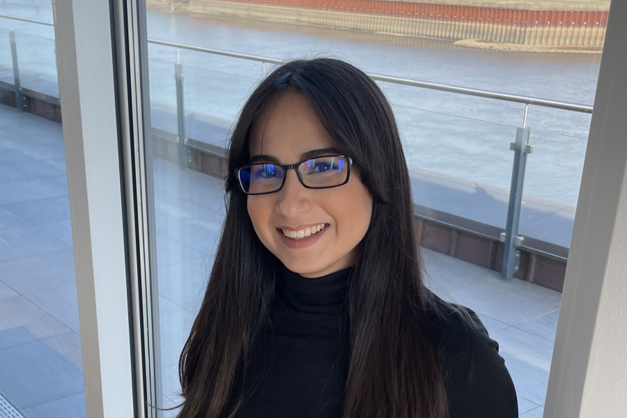 HR-Mitarbeiterin Hatice Erkekoğulları. Sie hat lange, dunkle Haare und einen schwarzen Rollkragenpullover an. Sie steht vor einem großen Fenster mit Blick auf den Fluss Weser und der Stadt Bremen im Hintergrund. 