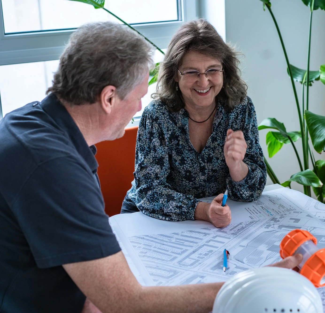 Zwei Mitarbeitende besprechen gemeinsam Baupläne an einem Tisch. Im Vordergrund liegen Schutzhelme und orange Rohrverbinder.