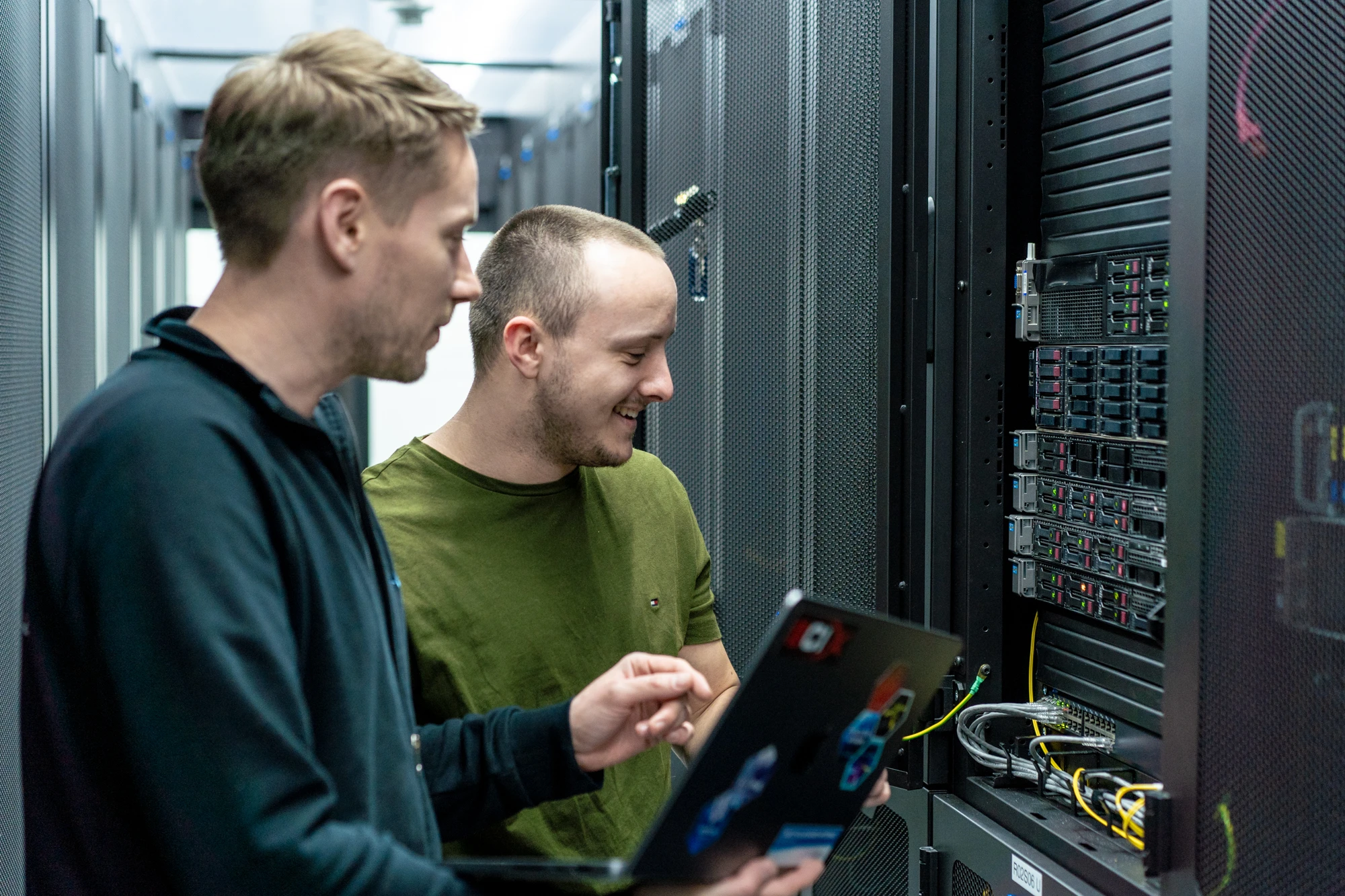 Zwei Mitarbeitende prüfen gemeinsam die Netzwerktechnik an einem geöffneten Serverschrank im Rechenzentrum. Einer von ihnen hält einen Laptop.
