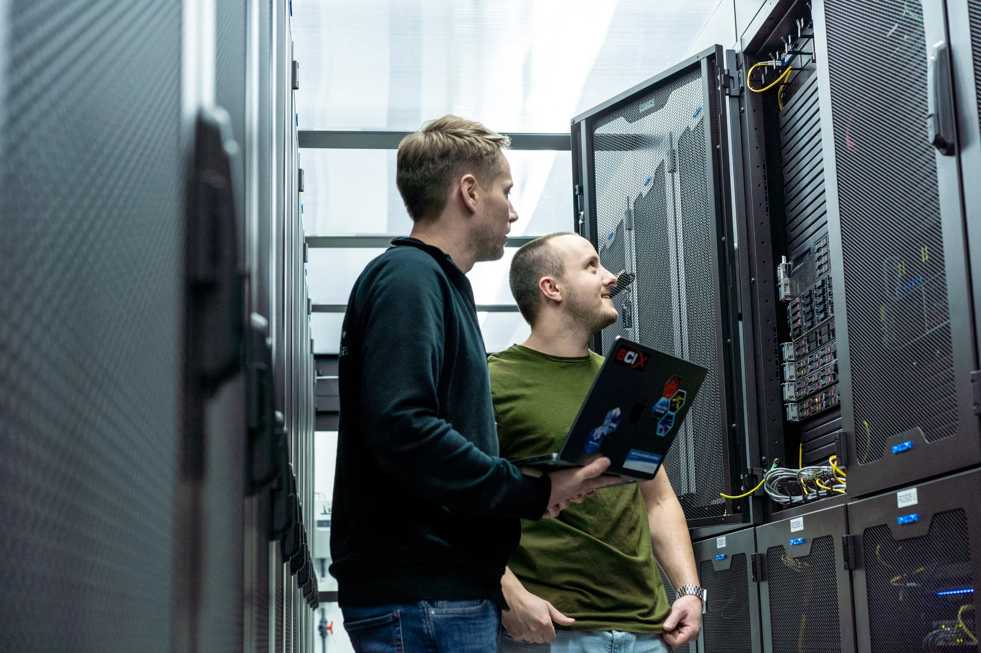 Zwei Mitarbeitende prüfen gemeinsam die Netzwerktechnik an einem geöffneten Serverschrank in einem Rechenzentrum. Einer von ihnen hält einen Laptop.