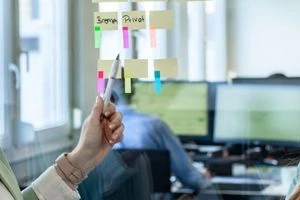 Eine Scheibe mit Klebezetteln, auf die jemand mit einem Stift zeigt. Im Hintergrund sitzt jemand an einem Schreibtisch.